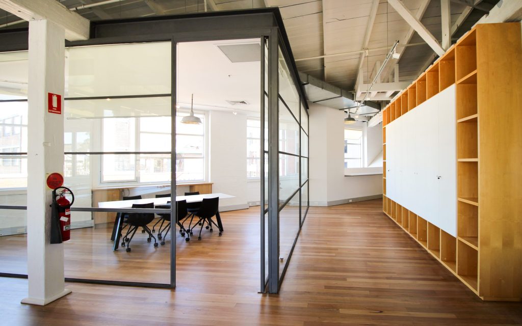 An office area with a glass partitioned meeting room in the left and a hallway beside it with wall cabinets/shelves to the right